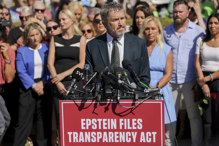 Un homme en costume, entouré d’une foule, se tient devant un pupitre recouvert de microphones sur lequel est inscrit « EPSTEIN FILES TRANSPARENCY ACT » (loi sur la transparence des dossiers Epstein)
