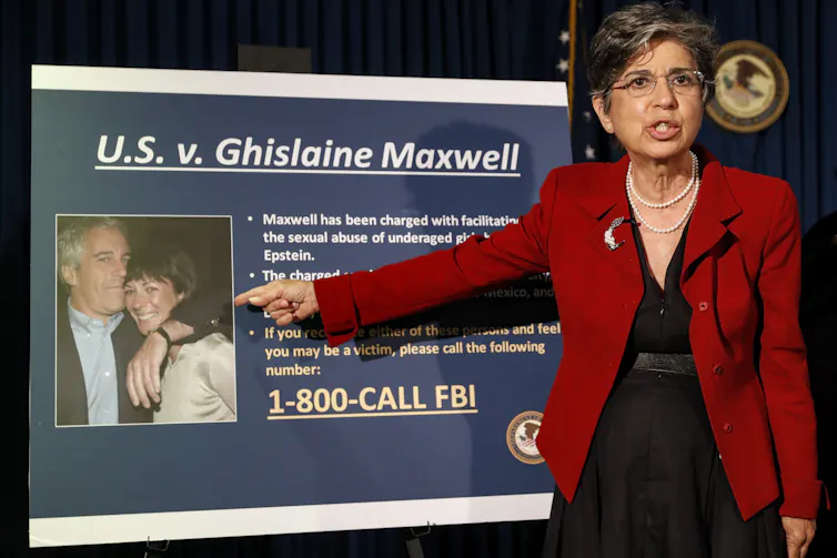 A woman in a red blazer points to a poster showing a man hugging a woman.