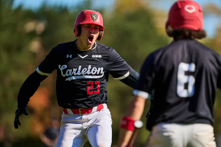 A baseball player in a black jersey that says Carleton celebrates with his arms thrown back