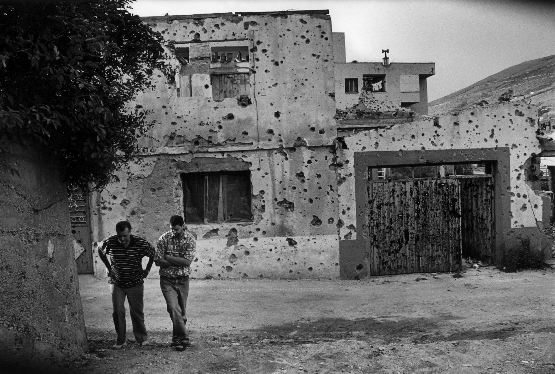 Two people walk down a street in front of a damaged building.