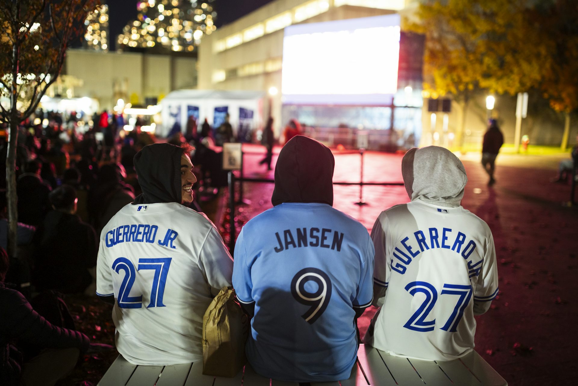 Seated fans wearing blue jays baseball jerseys seen from the back looking towards a screen.