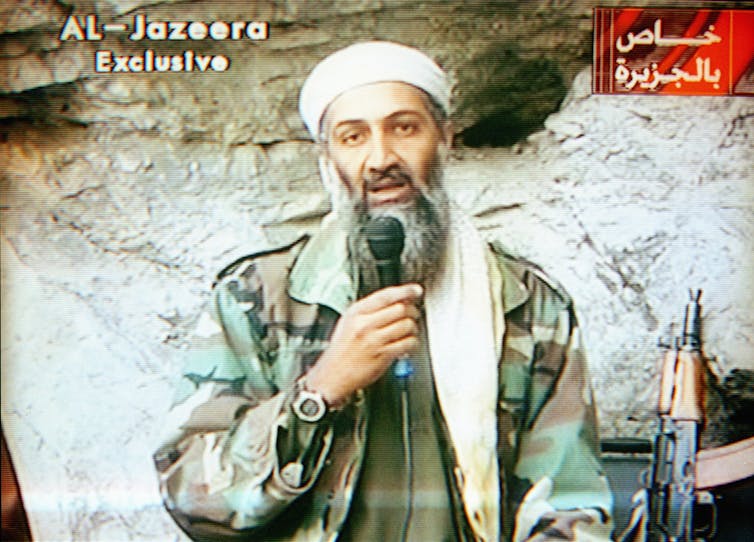 A man with a grey beard and turban speaks into a microphone on TV.