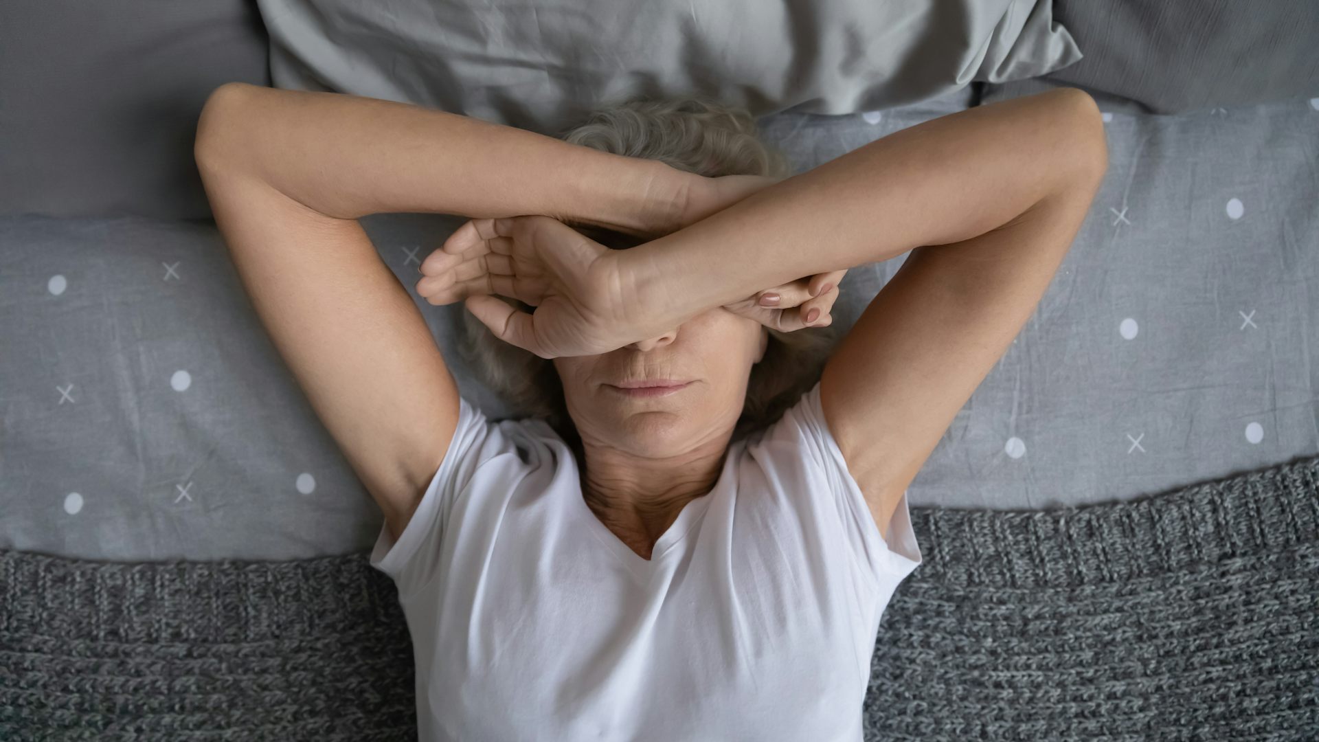 Woman lying on grey bedsheets with her arms over her face.