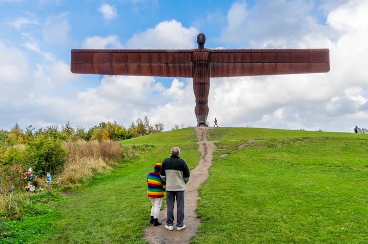 angel of the north big steel sculpture on green grass, blue sky with two people walking towards it along path