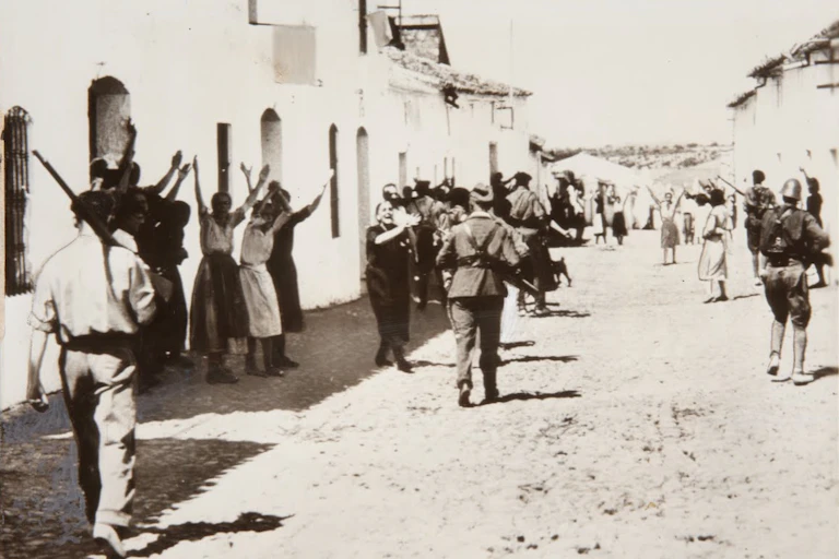 Foto en blanco y negro con mujeres levantando los brazos al paso de hombres armados