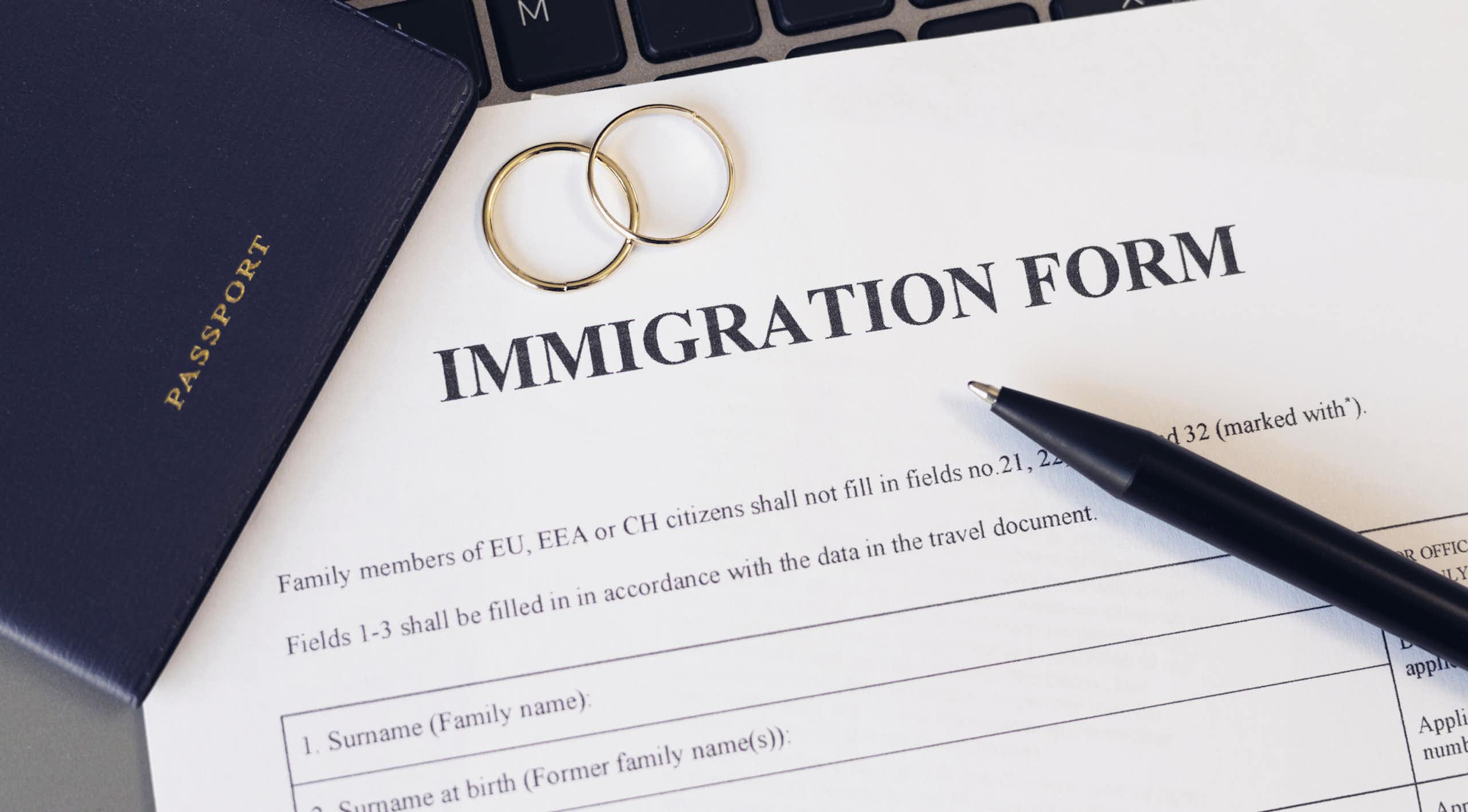 A white form that says 'immigration form' at the top rests near a passport, a pen and two gold rings.