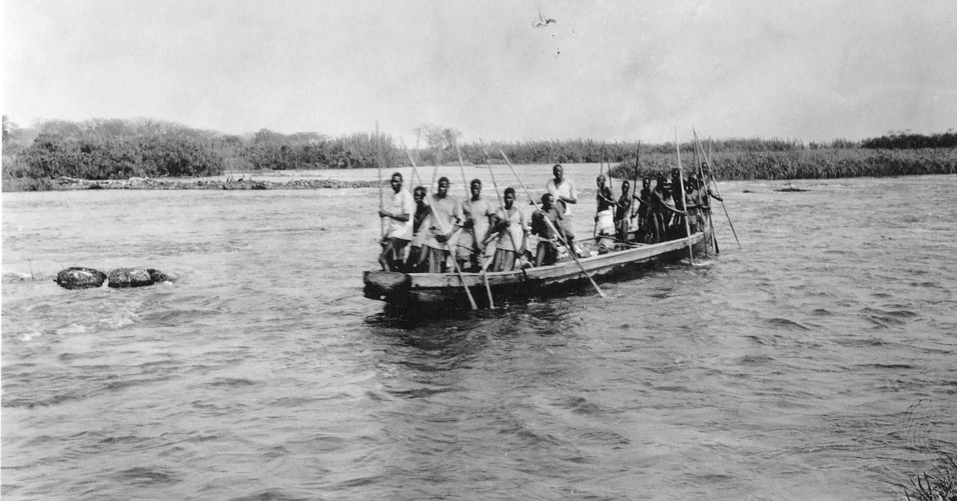 A black and white photo of a boat on a wide river with a crowded group of Africans holding long sticks.