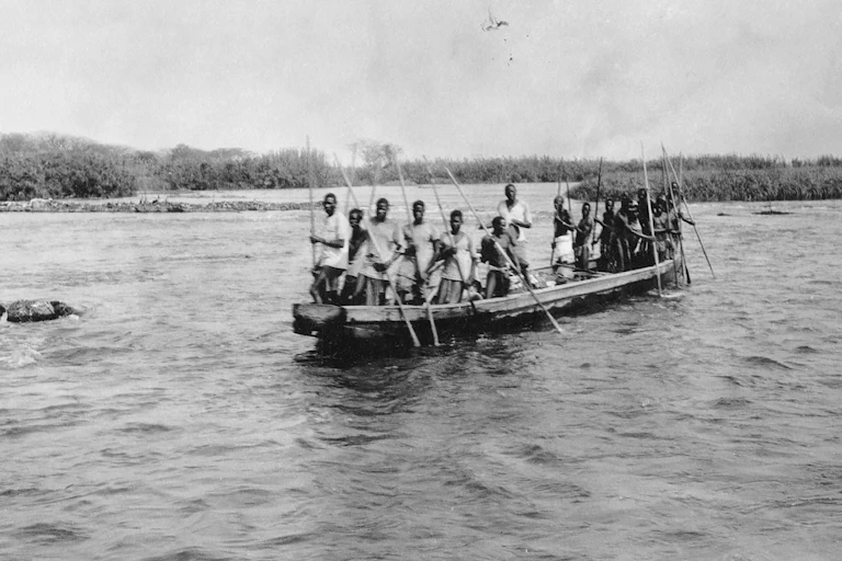 A black and white photo of a boat on a wide river with a crowded group of Africans holding long sticks.