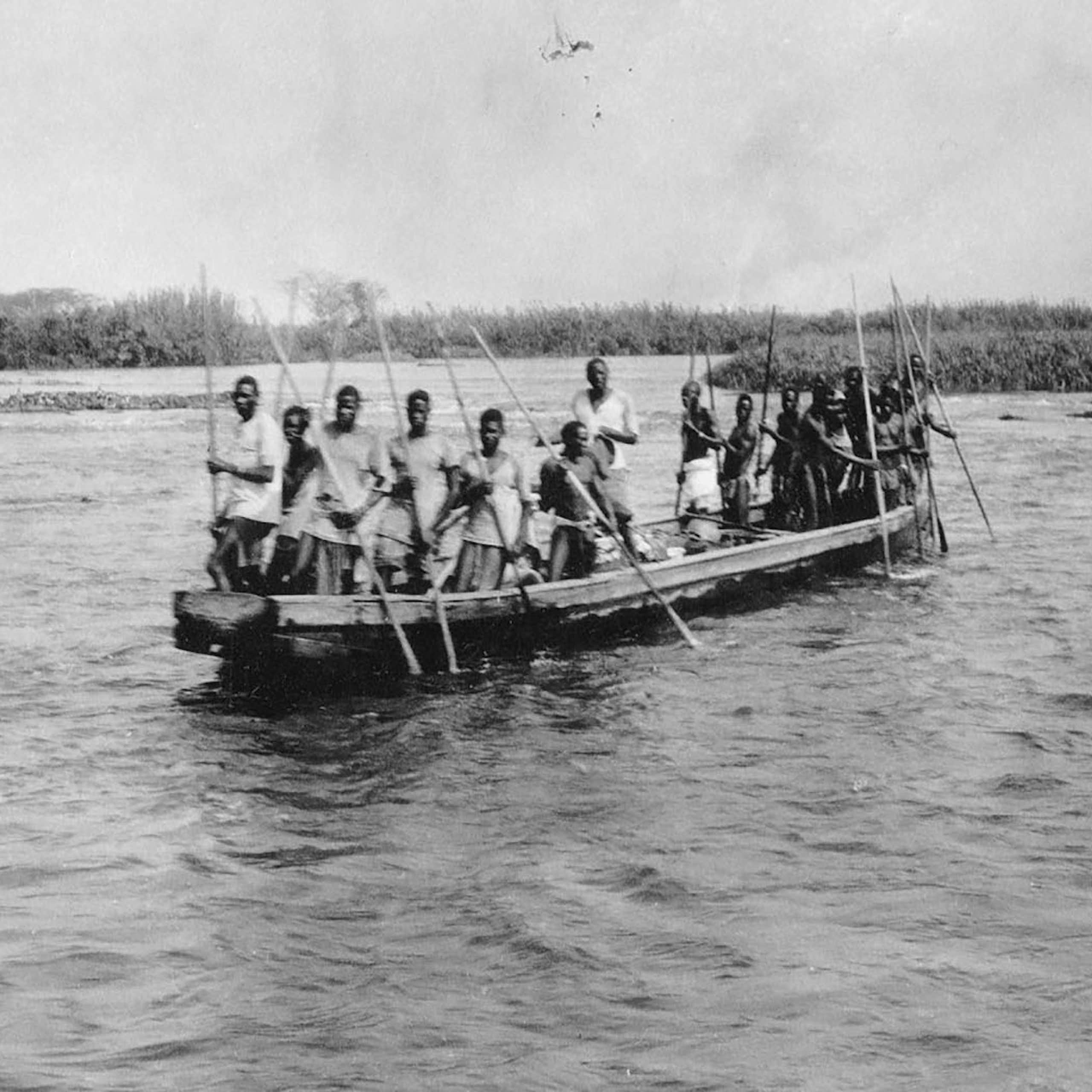 A black and white photo of a boat on a wide river with a crowded group of Africans holding long sticks.