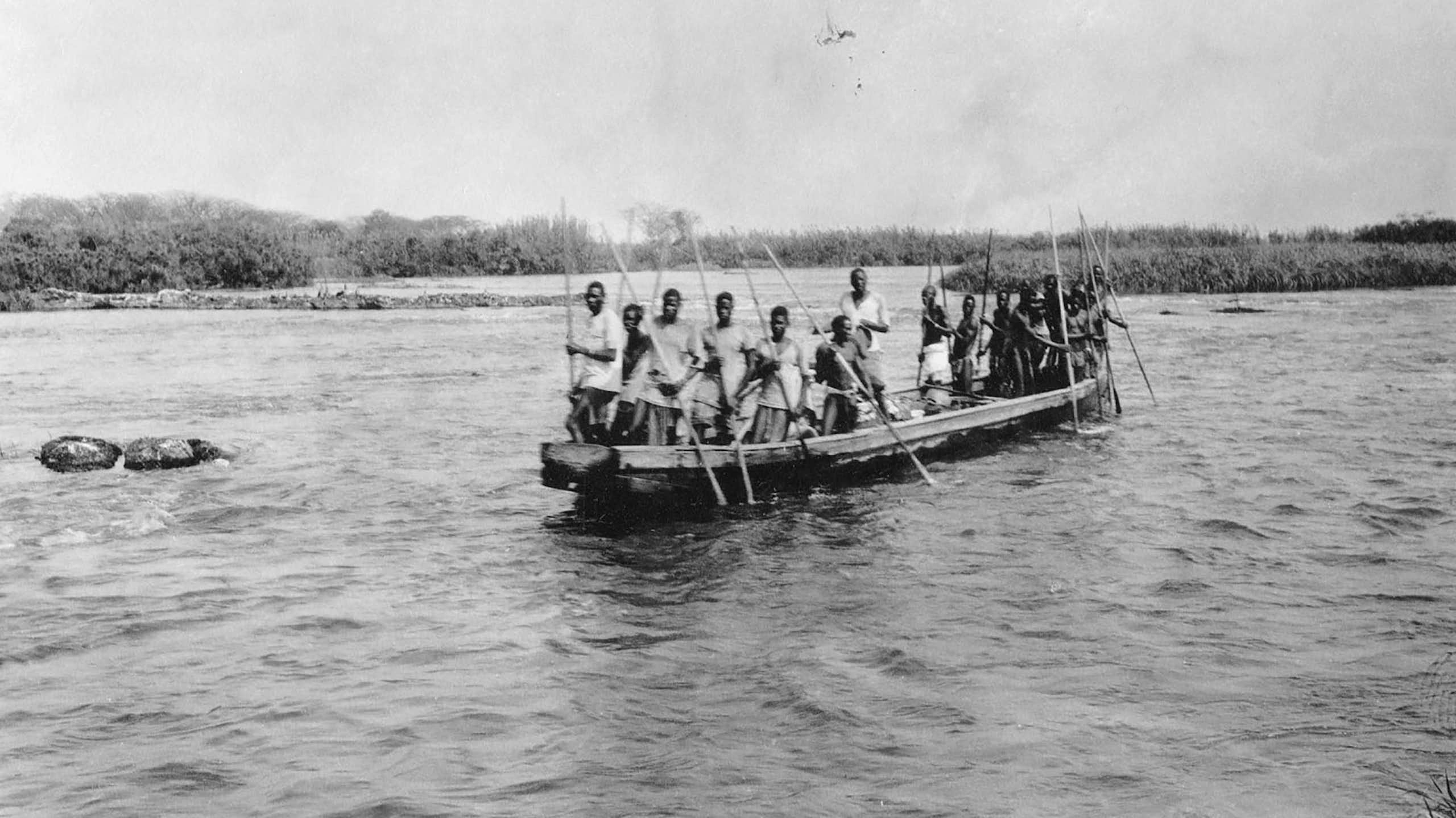 A black and white photo of a boat on a wide river with a crowded group of Africans holding long sticks.