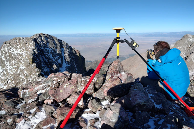 A man squats next to a tripod on the top of a mountain.