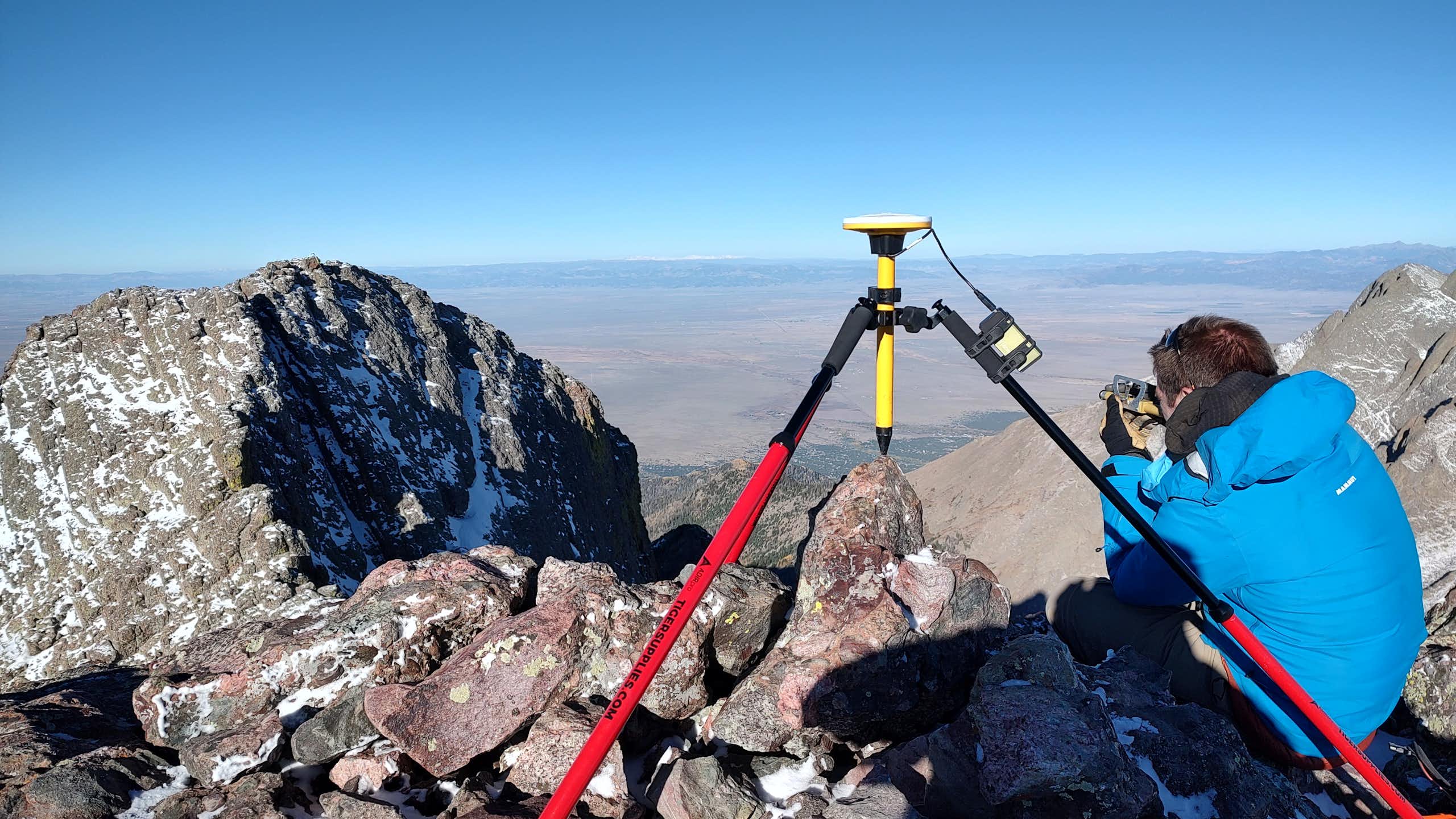 A man squats next to a tripod on the top of a mountain.