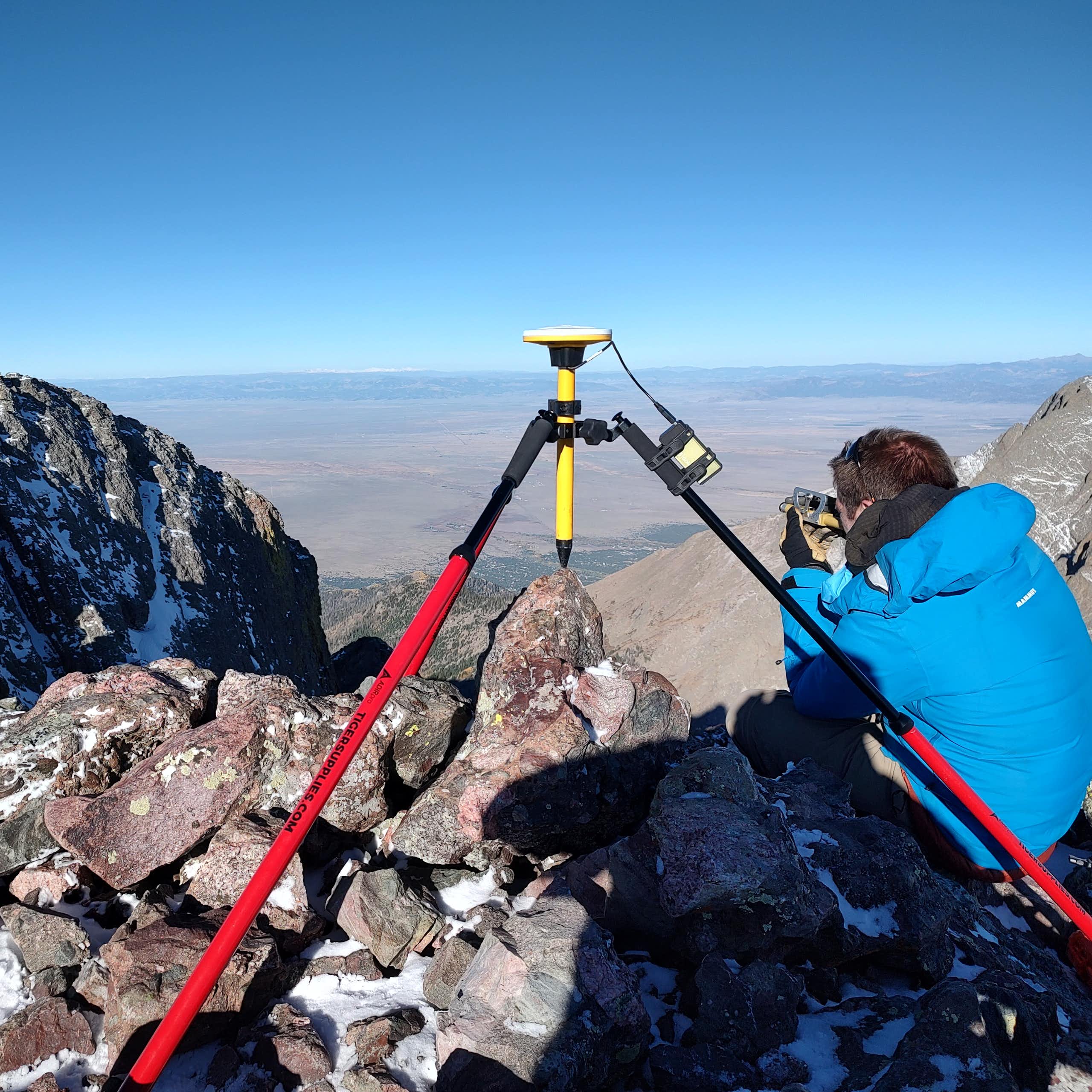 A man squats next to a tripod on the top of a mountain.