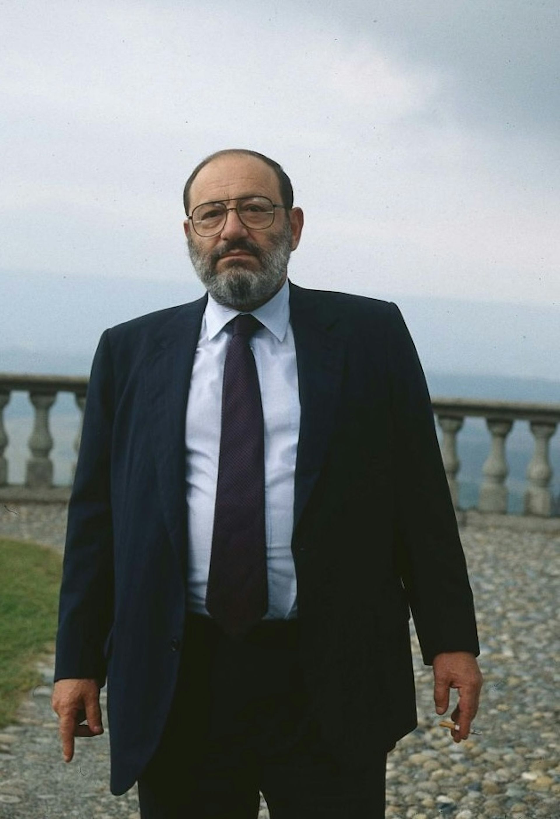 An older man with a beard, glasses and a suit poses while holding a cigarette.