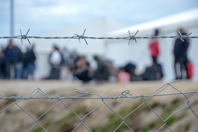 Barbed wire at a refugee camp, with people out of focus in the background
