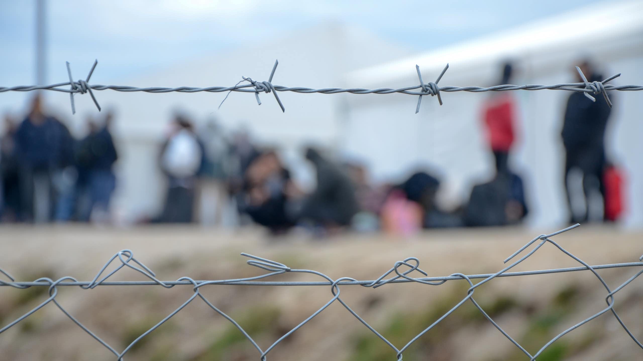 Barbed wire at a refugee camp, with people out of focus in the background