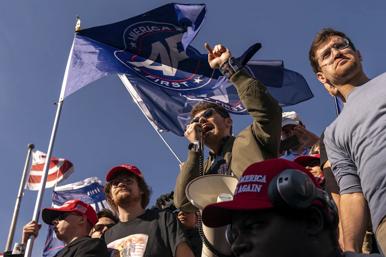 Young man at a rally speaking into a microphone, surrounded by other young men.