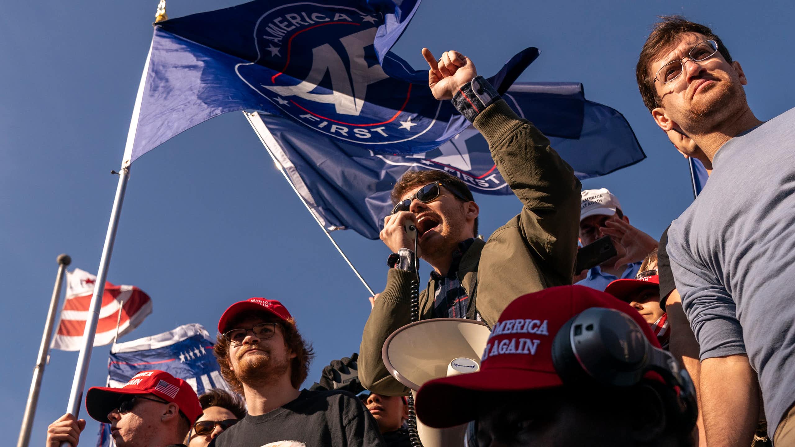 Young man at a rally speaking into a microphone, surrounded by other young men.