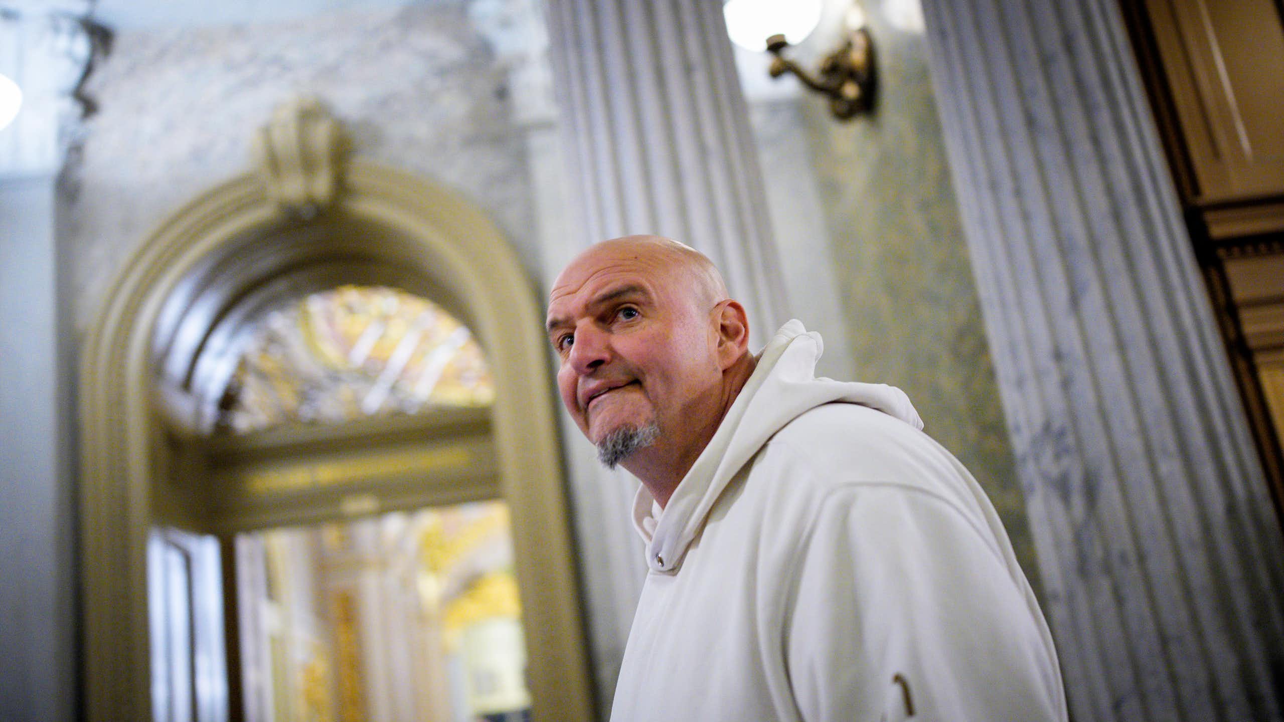 Large bald man wearing white hoodie walks past Grecian columns on ornate room