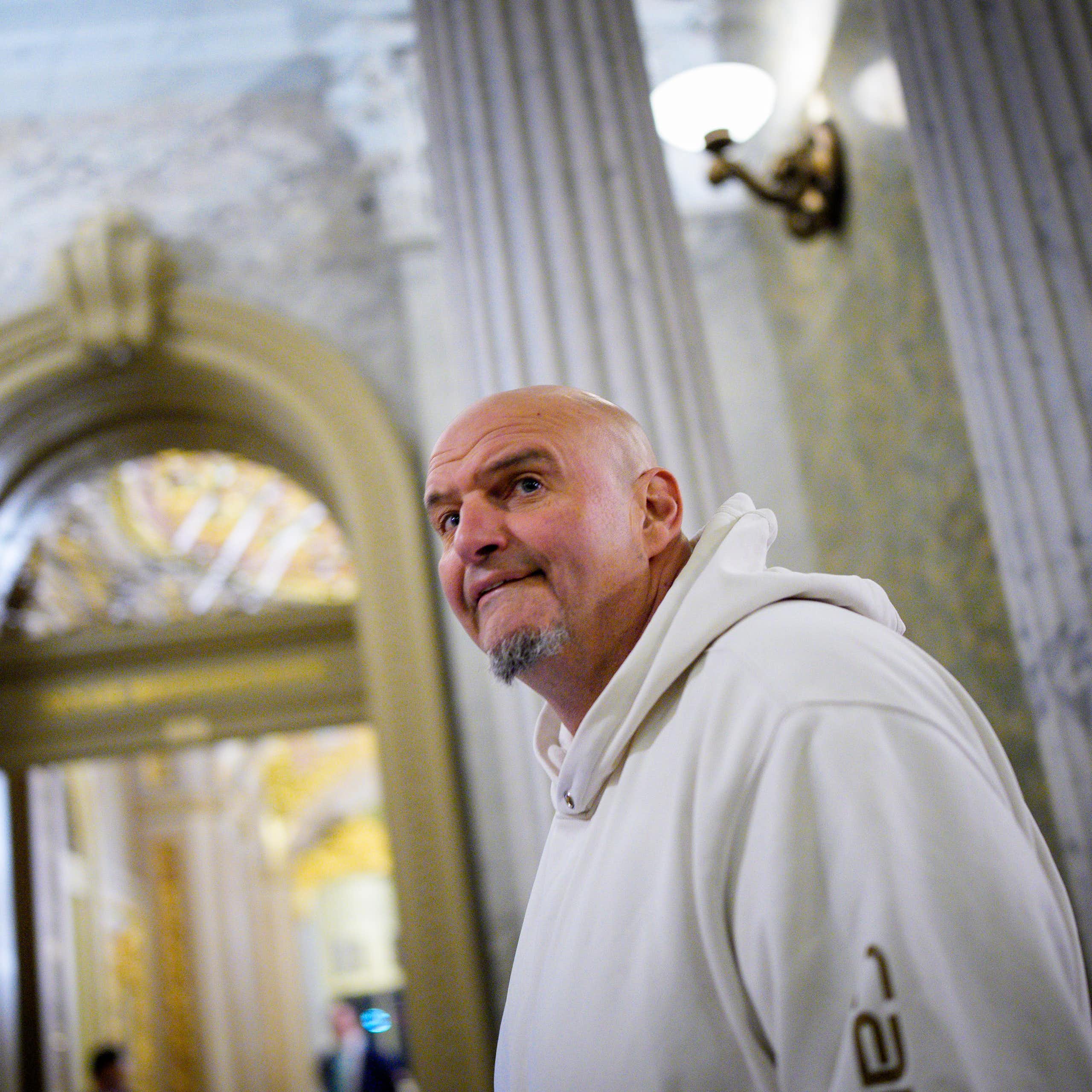 Large bald man wearing white hoodie walks past Grecian columns on ornate room