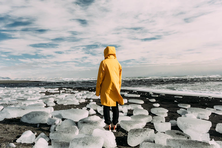 Person is yellow raincoat facing away from camera, standing on a rocky beach covered with lumps of ice under a cloudy sky.