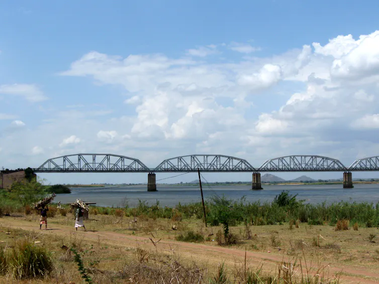 Women walk on a dirt road, behind them a river with a large bridge.