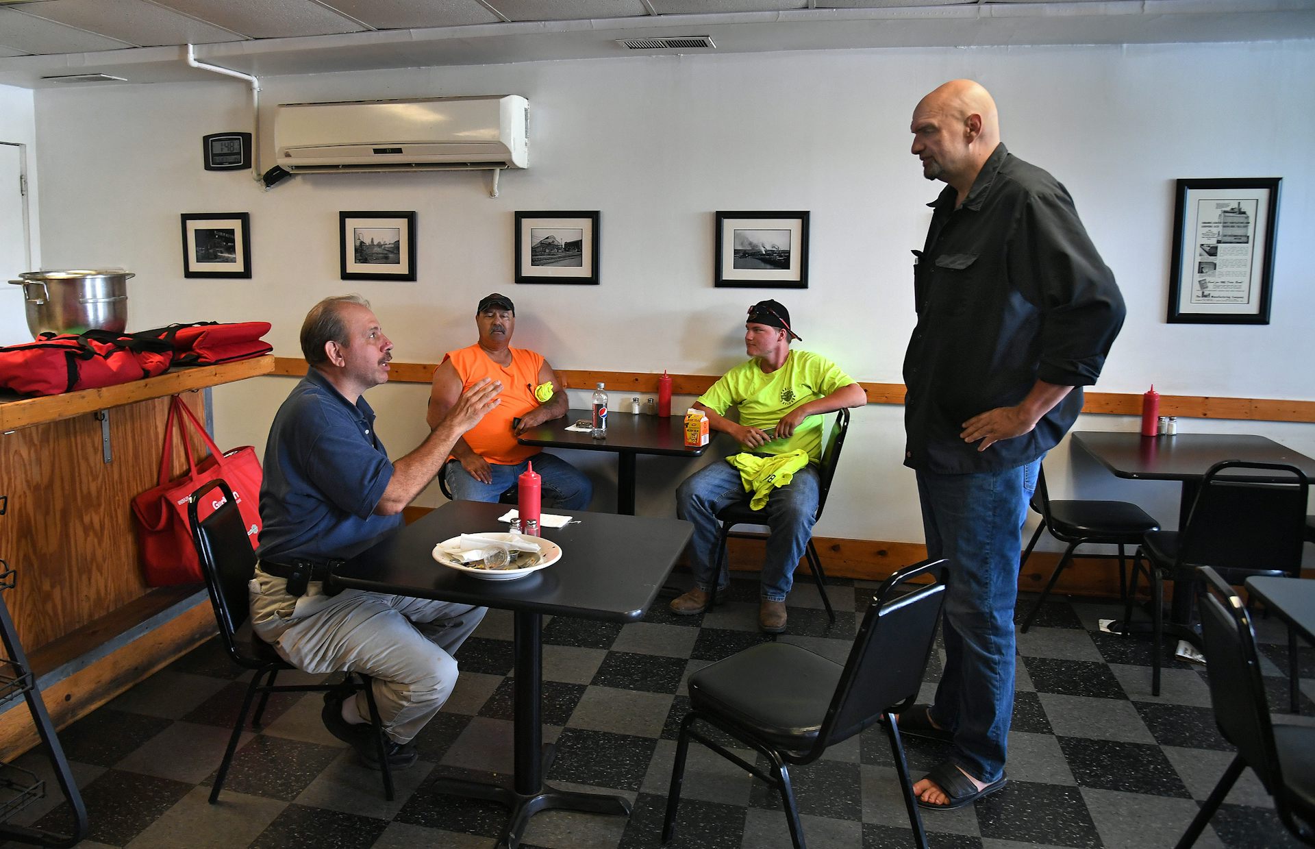Tall bald man in black shirt and jeans speaks to three workers sitting in a diner