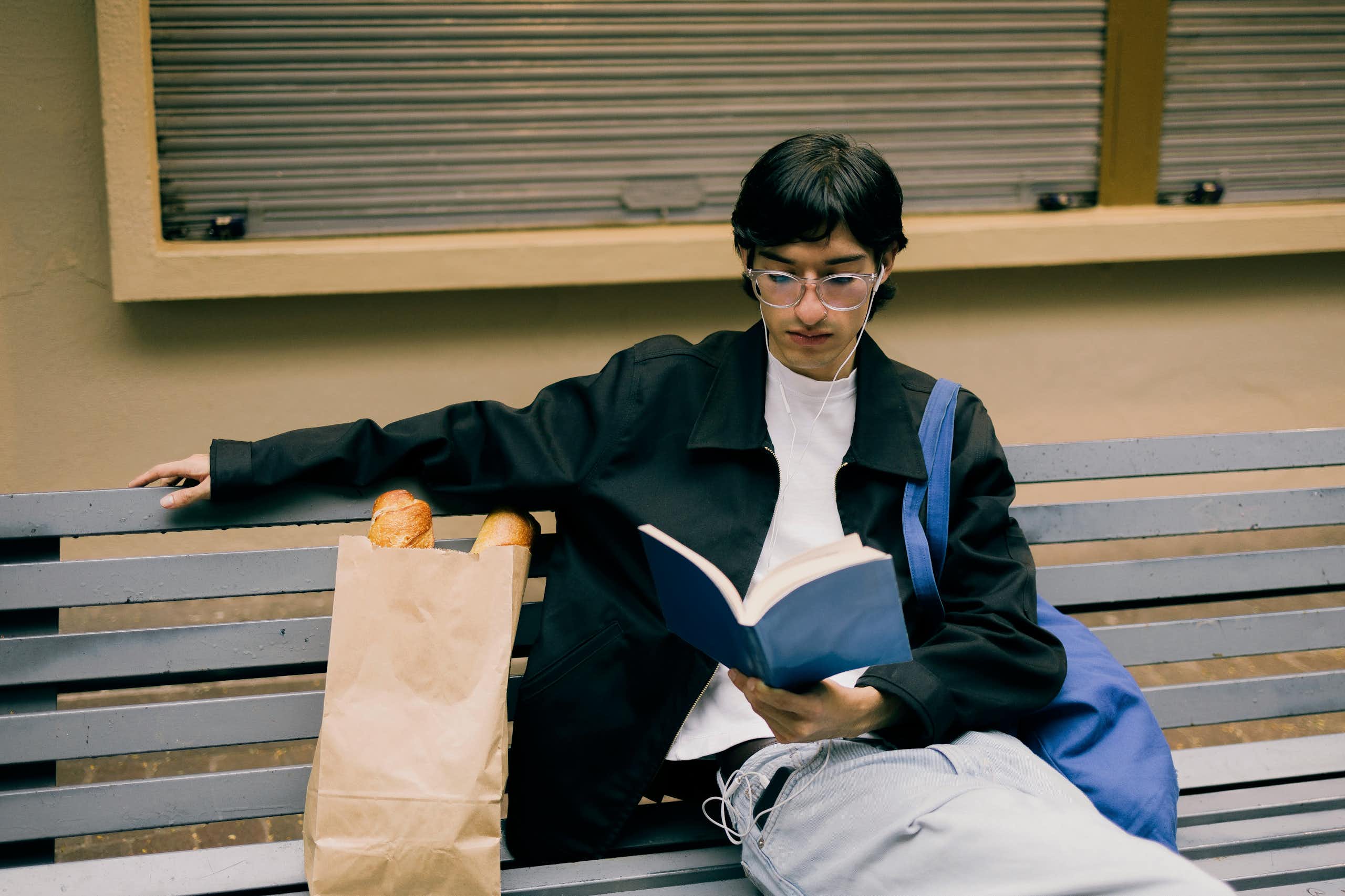 Un jeune homme lisant un livre sur un banc, avec un sac fourre-tout porté à l’épaule.