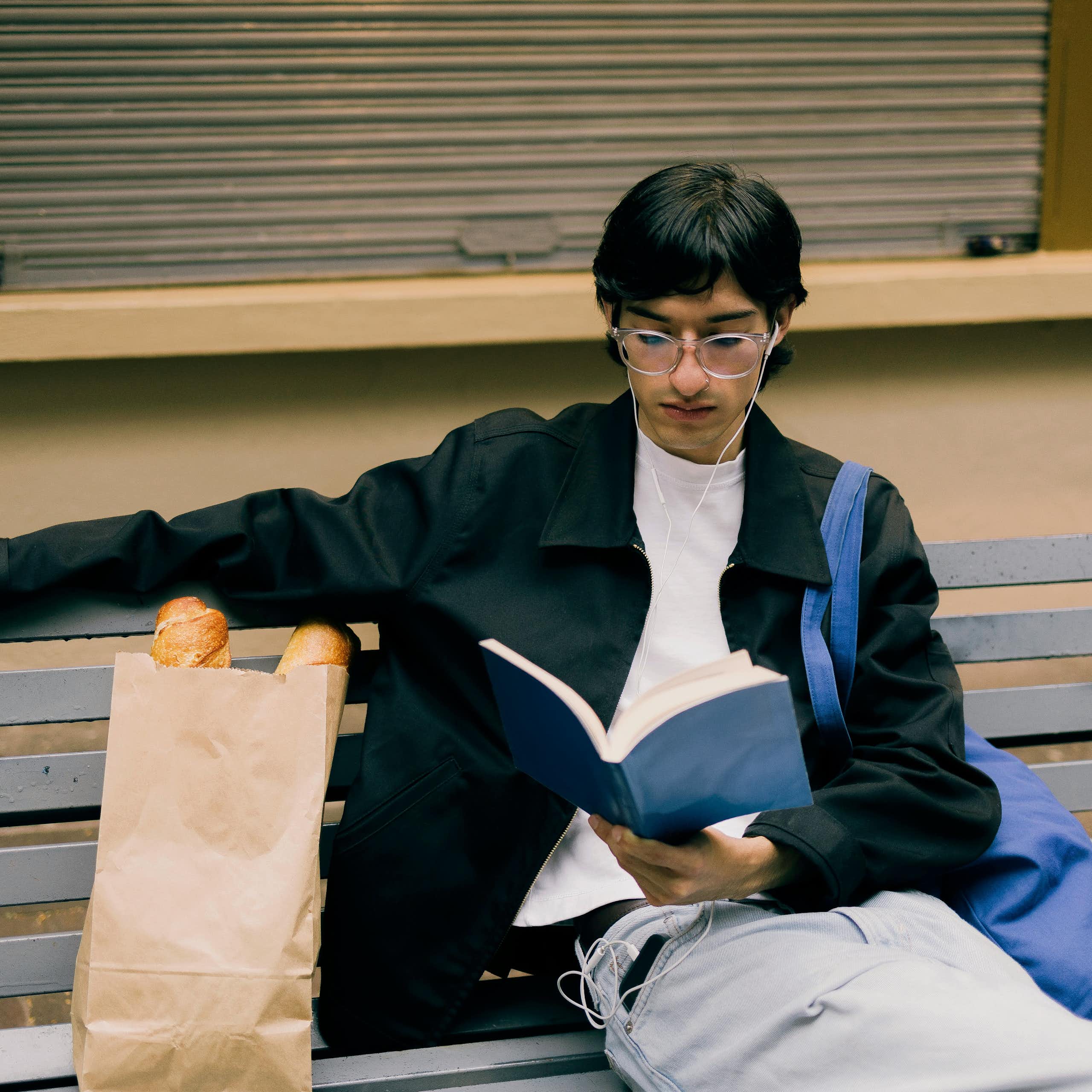 Un jeune homme lisant un livre sur un banc, avec un sac fourre-tout porté à l’épaule.