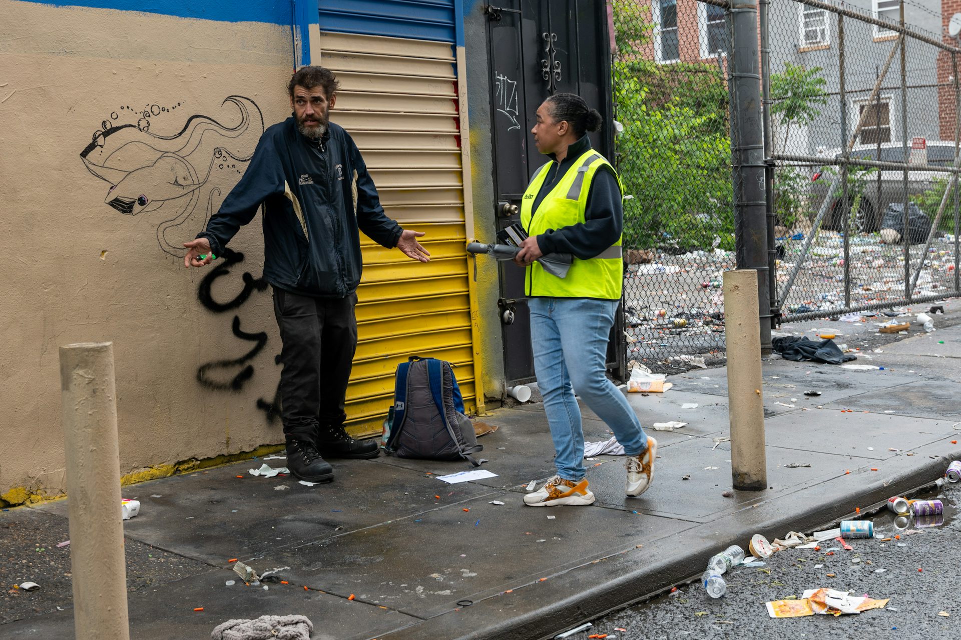 Woman in fluorescent yellow vest speaks with disheveled man standing on trash-strewn sidewalk