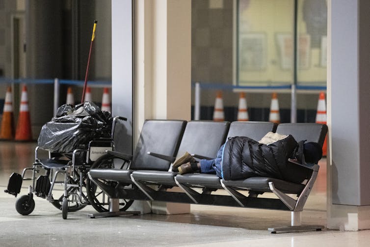 Person sleeps on chairs with wheelchair nearby
