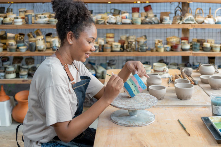A young and happy woman sitting in a pottery shop making a cup and painting it