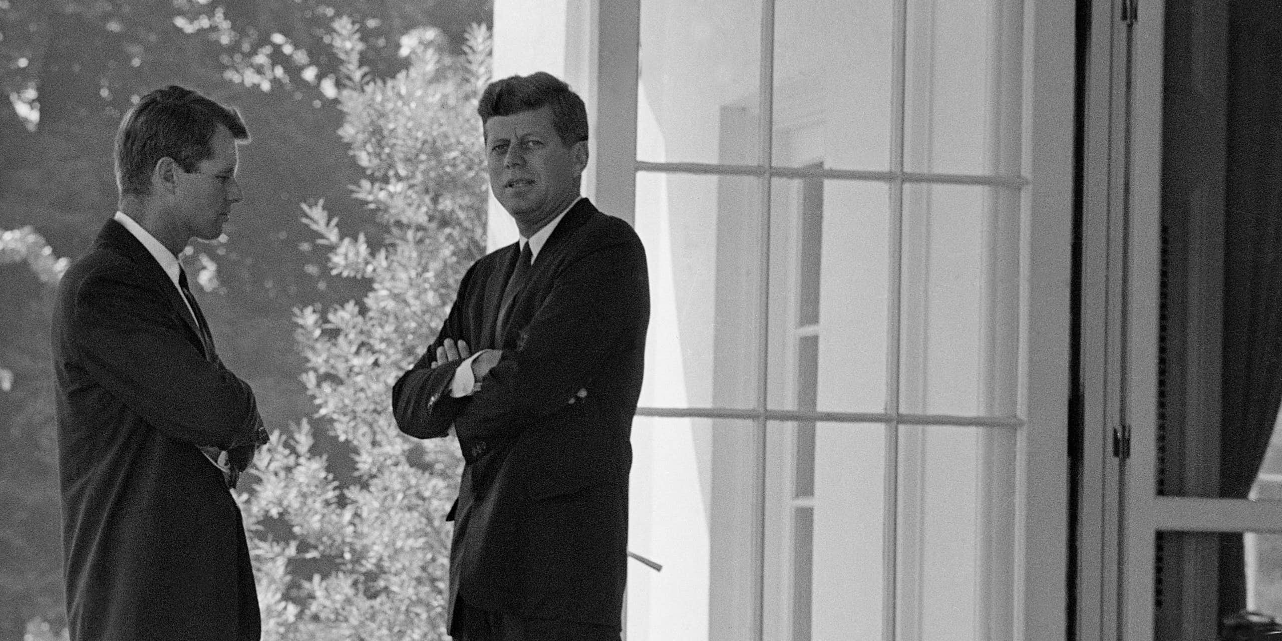 Two men in suits standing outside a room with a large glass door open behind them.