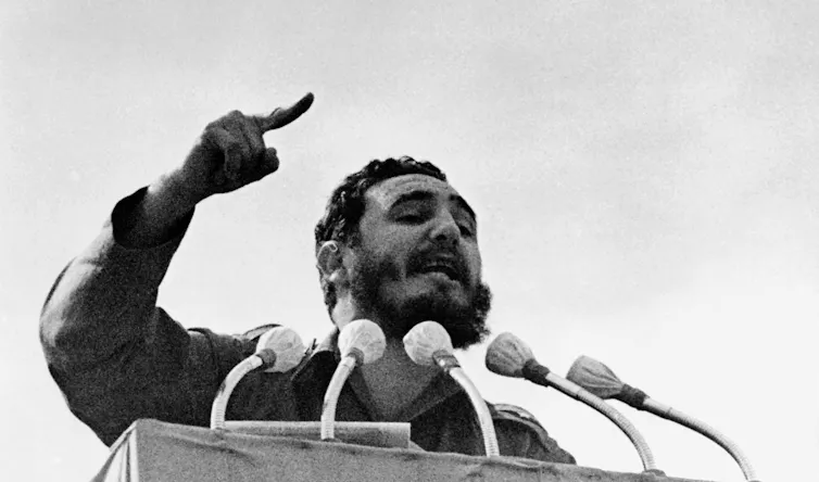 A bearded man at a lectern with a raised arm and speaking into multiple microphones.