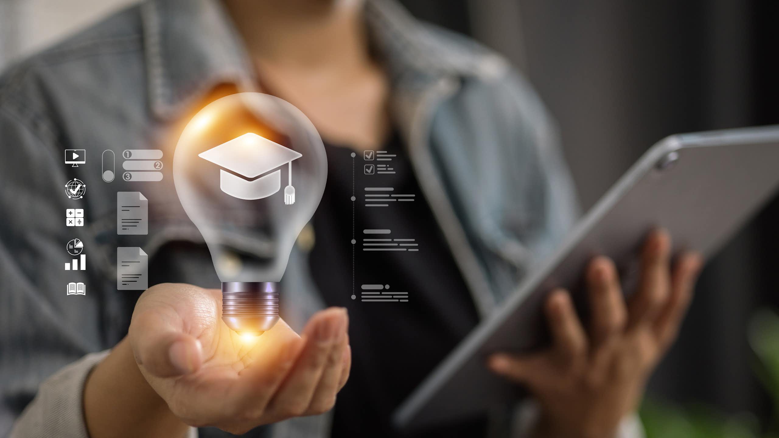 A person wears a grey blazer and hold up an illuminated lightbulb with a graduation cap inside of it, and a tablet in another hand.