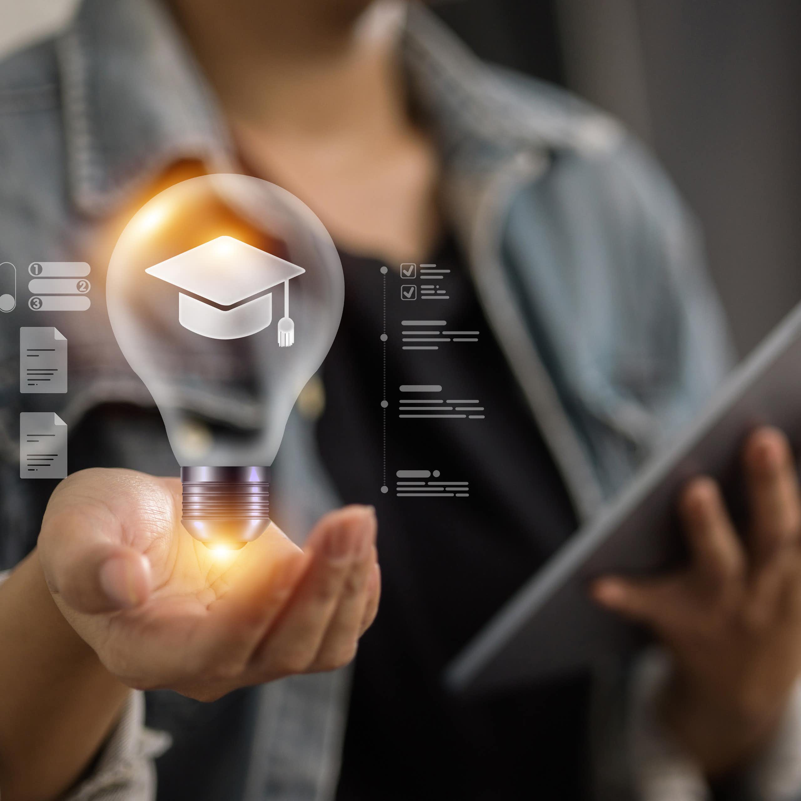 A person wears a grey blazer and hold up an illuminated lightbulb with a graduation cap inside of it, and a tablet in another hand.