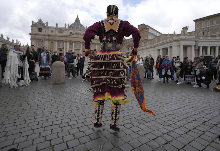 A person in a jingle dress in front of a domed building while others in a circle watch.