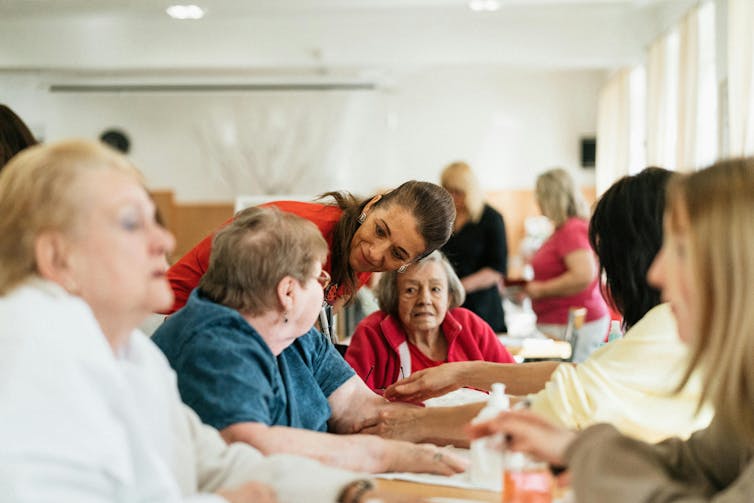 Las personas mayores están sentadas en una mesa y una persona más joven está de pie y les habla.