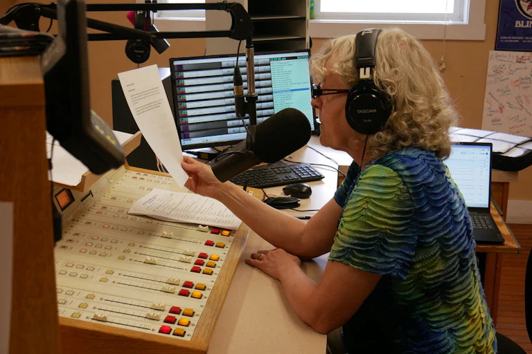 A woman speaks into a large microphone while sitting at a desk with a lot of recording equipment.