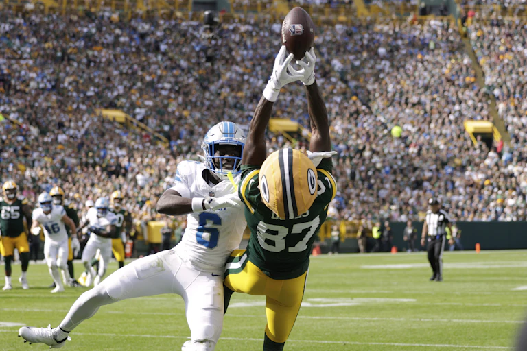 A player in a green and gold uniform falls backward while trying to catch a football.
