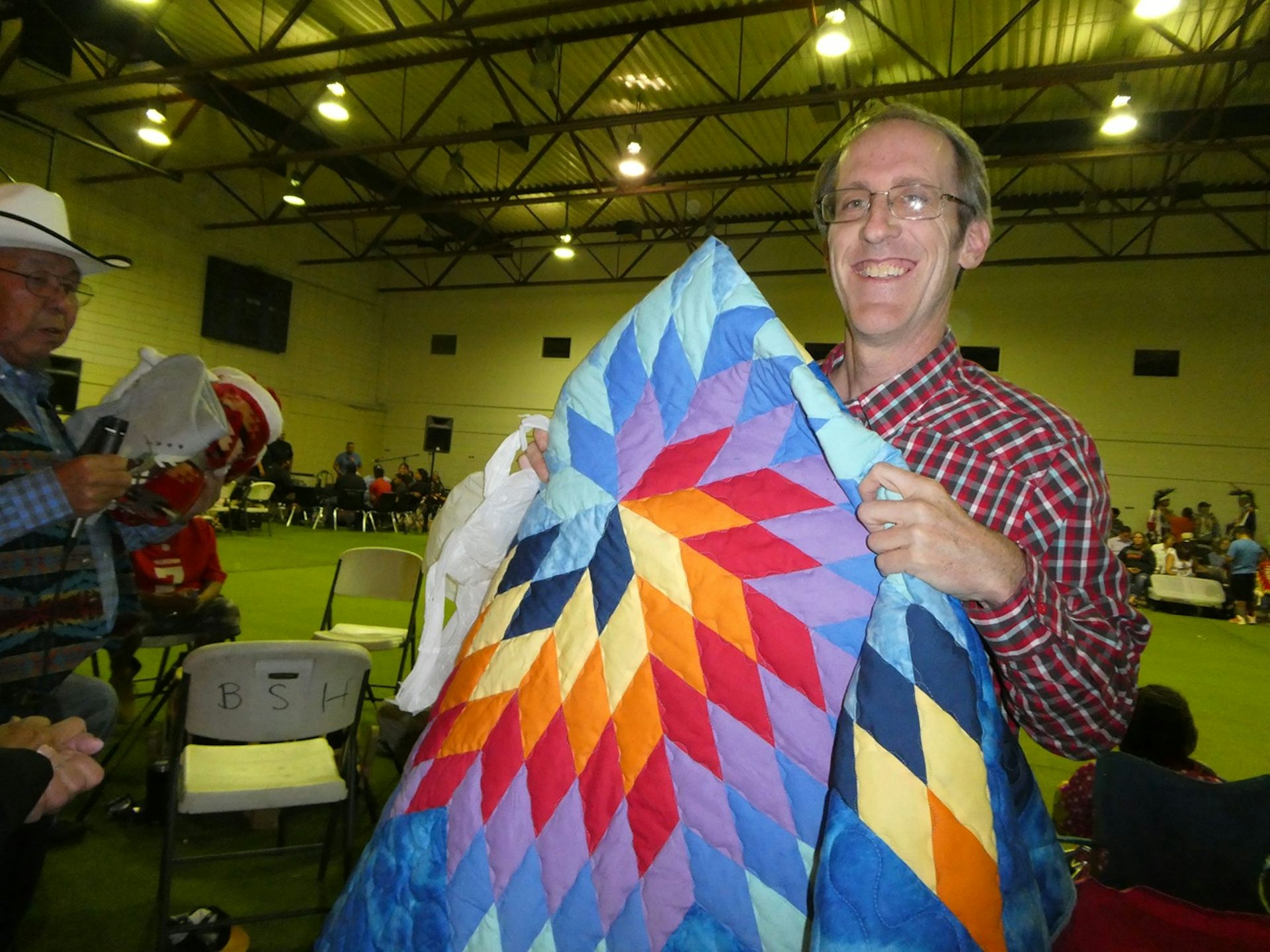 A man in a red gingham shirt holds a colorful quilted blanket.