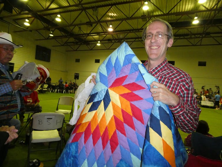 A man in a red gingham shirt holds a colorful quilted blanket.