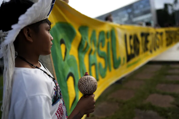 a child holding a maraca and wearing a headdress made of feathers stands near a large yellow banner at a demonstration