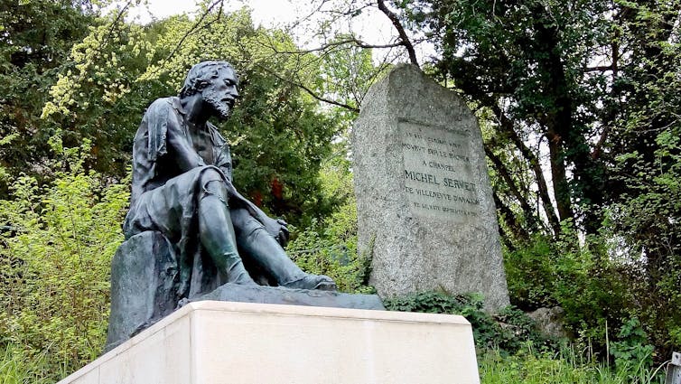 A dark-colored statue of a downcast, seated man, positioned in front of trees and bushes.