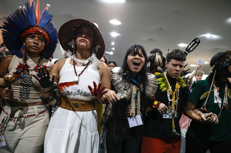 Uma fila de manifestantes de mãos dadas e gritando, usando joias indígenas