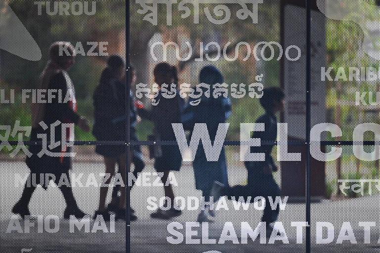 Children can be seen in the reflection of a window with 'welcome' written on it in various languages.