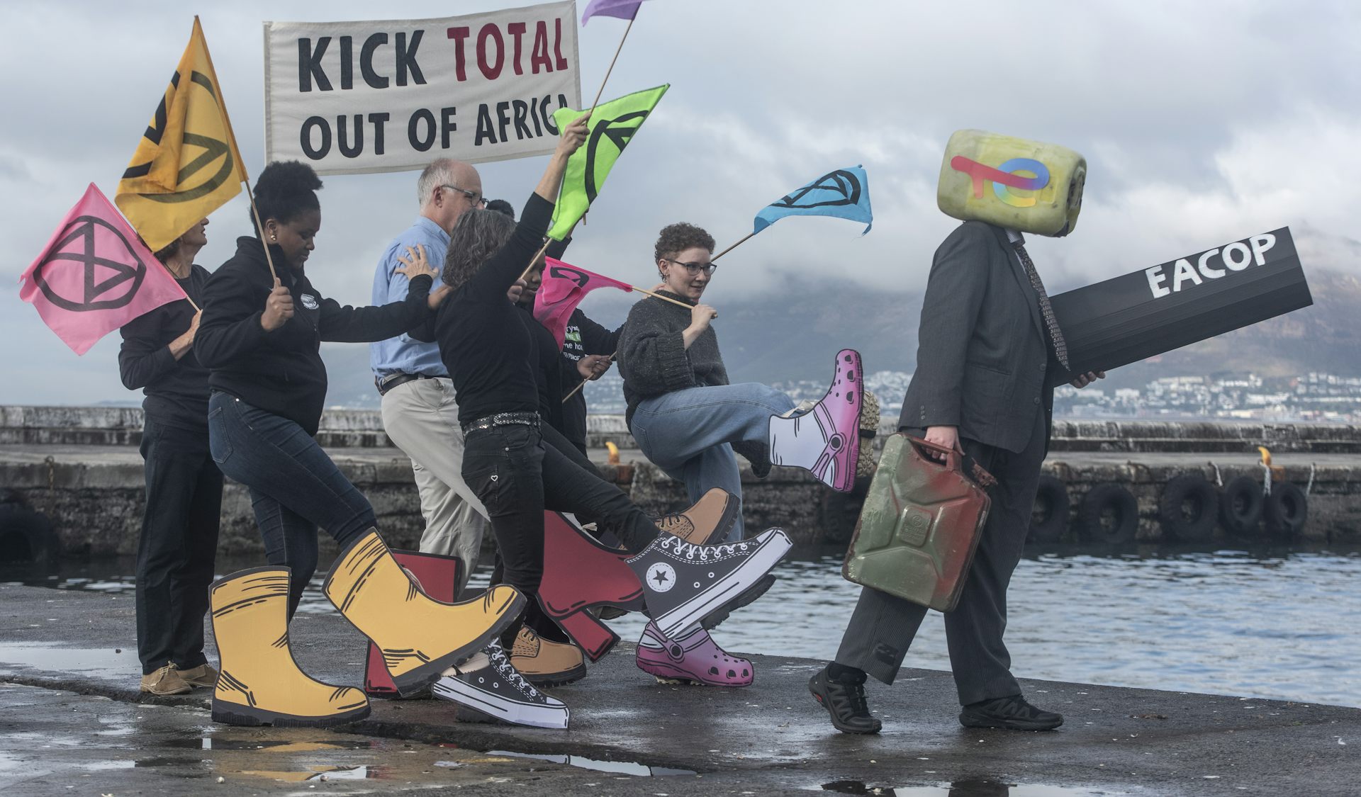 People holding a banner next to a harbour. The banner says "Kick Total out of Africa" and the people have giant cardboard shoes attached to their legs and are making kicking motions