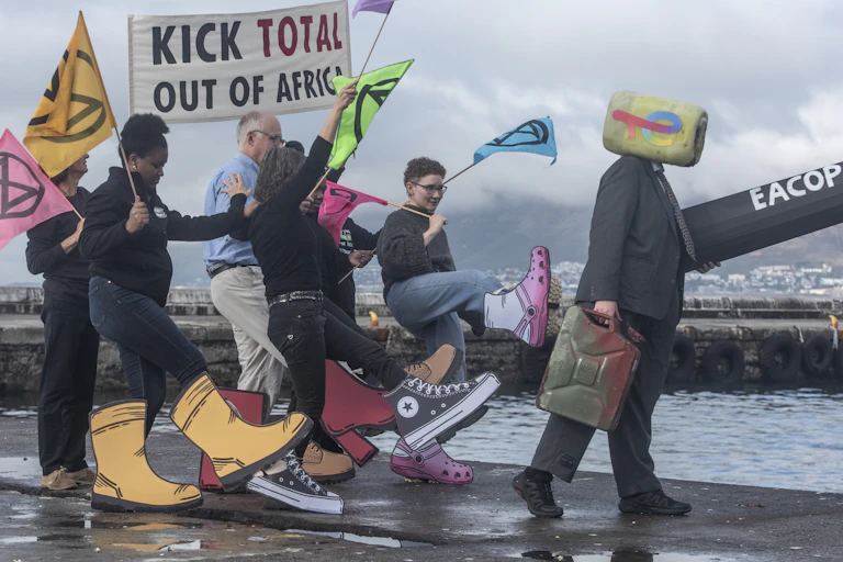 People holding a banner next to a harbour. The banner says "Kick Total out of Africa" and the people have giant cardboard shoes attached to their legs and are making kicking motions