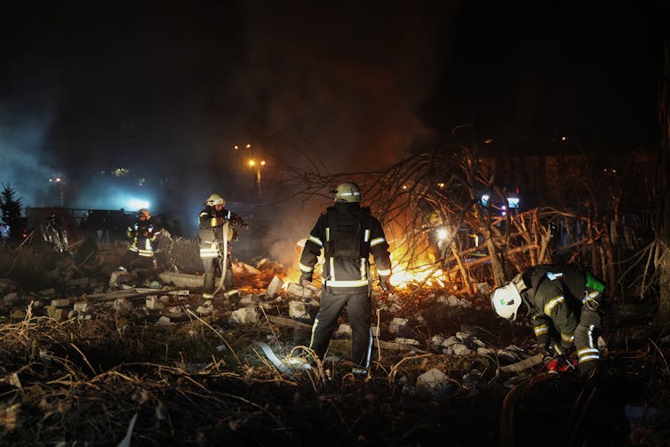 A rescue worker in a uniform stands in front of the rubble of a bombed building.