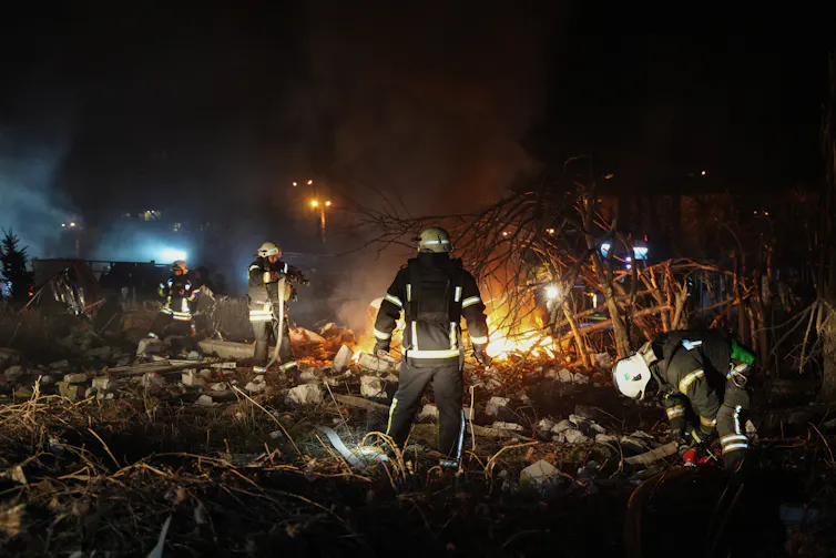A rescue worker in a uniform stands in front of the rubble of a bombed building.